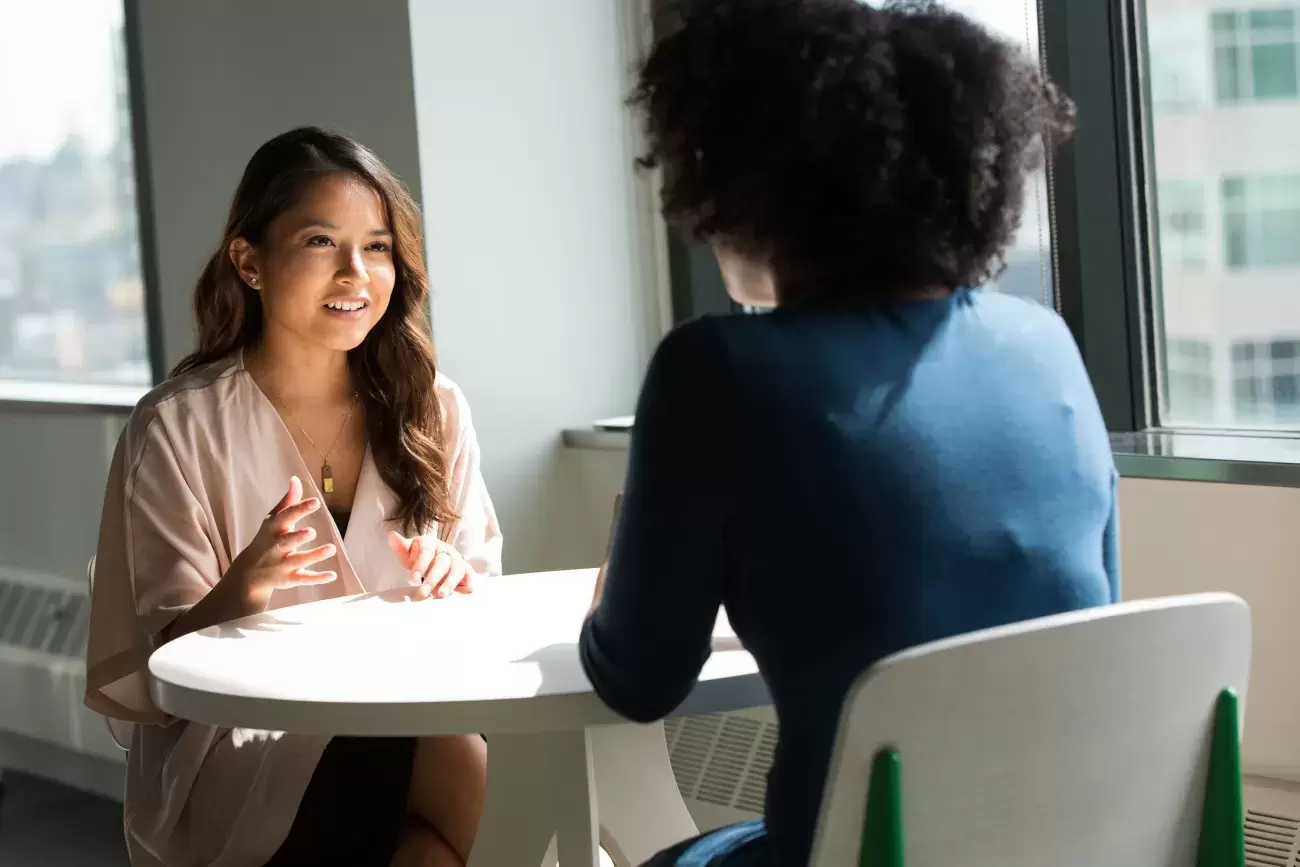 two people sit at table talking together, one with back to us