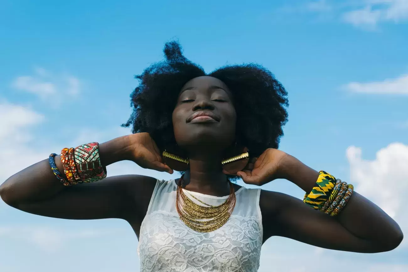 person wearing bangles and necklaces stands with only blue sky behind them, smiling peacefully with eyes closed