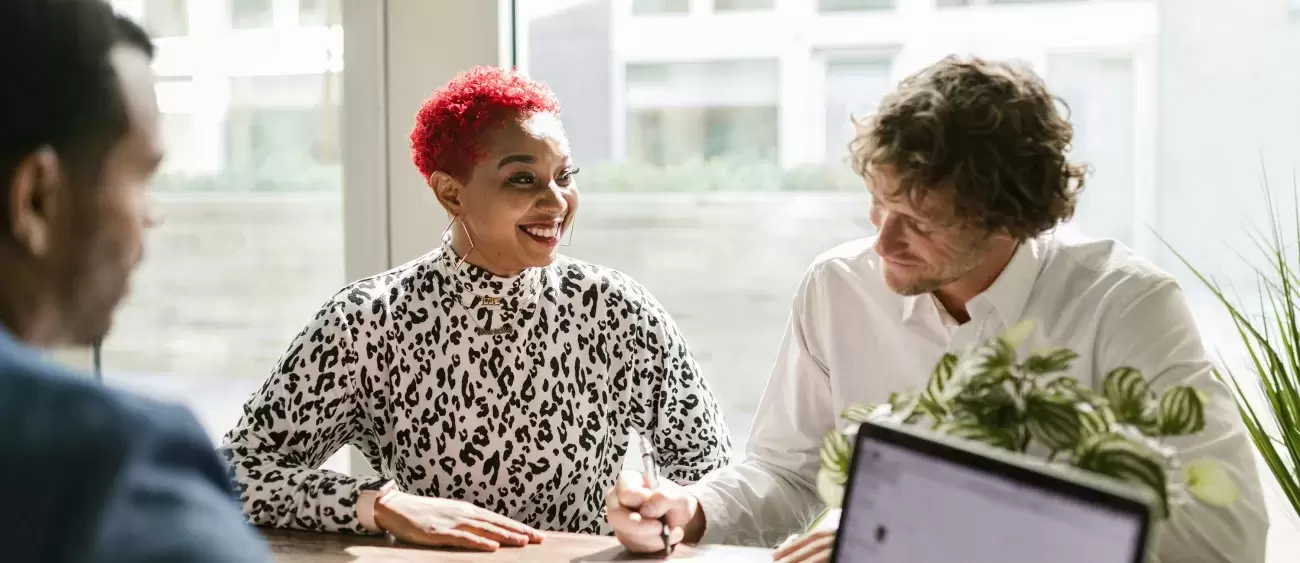 smiling people sit at a table looking at paperwork and computer together