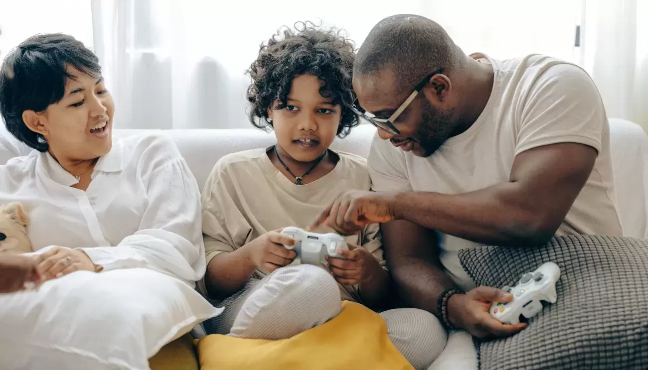 family sits on couch playing video games; dad leans over to show child how controller works