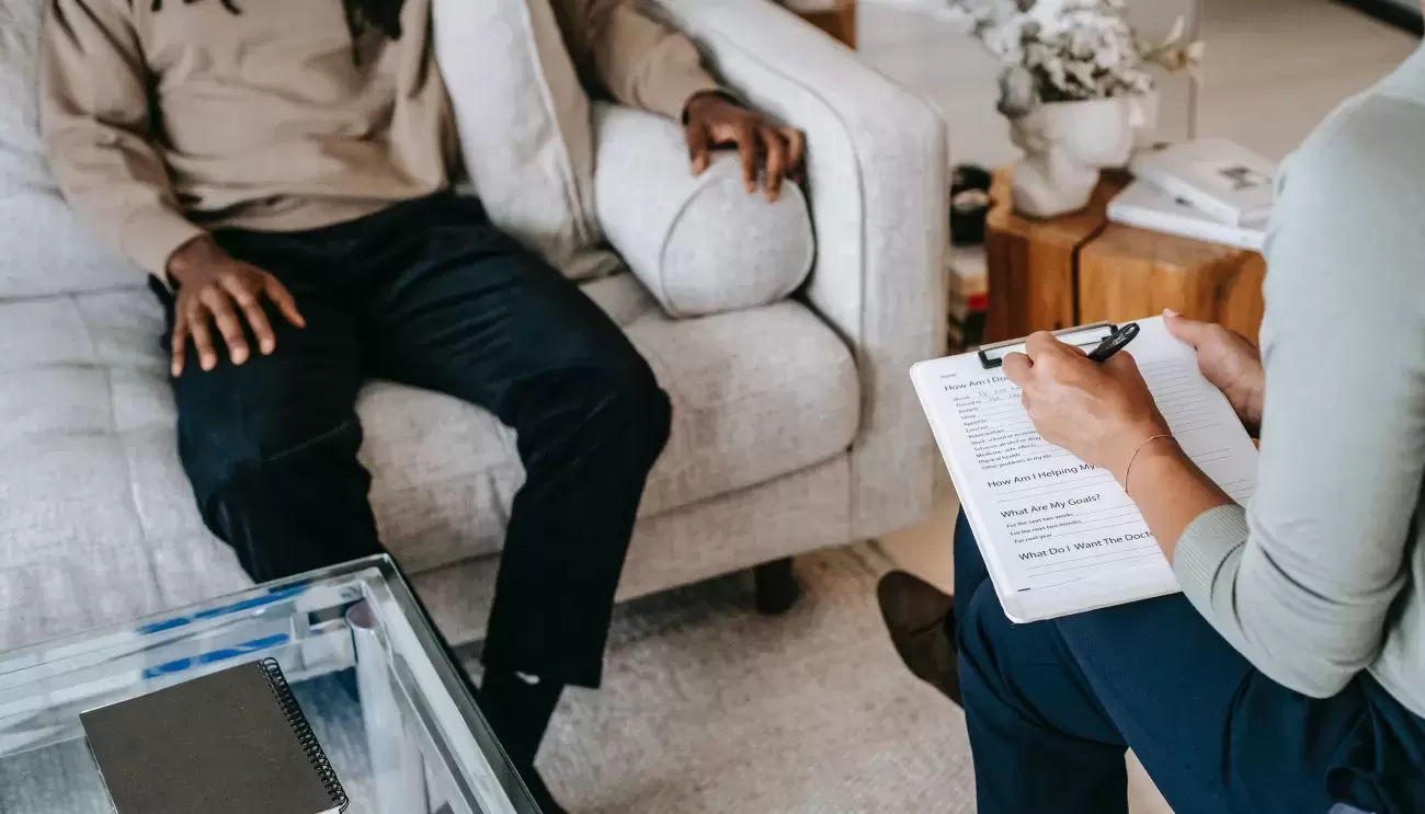 close-up of two people, one sitting on a couch with one hand on leg and one on the arm of the couch, other person sits in chair across from them and writes on notebook, table is on floor between them