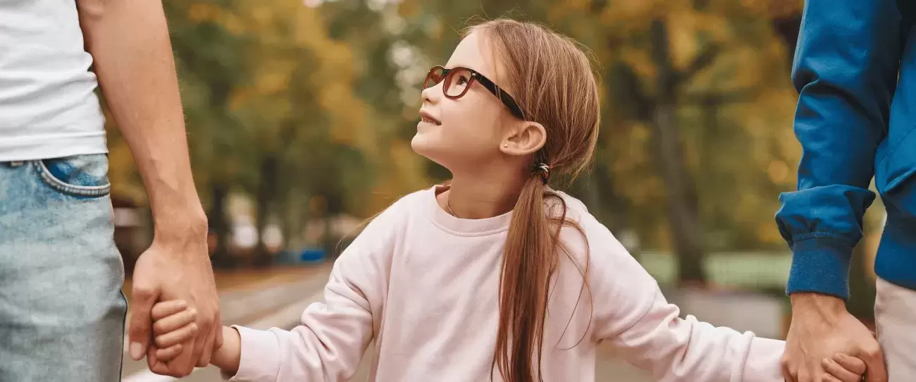 young child wearing glasses looks up while standing between and holding hands with two adults