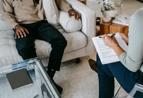 person sits on couch while another person sits across from them with clipboard and pen